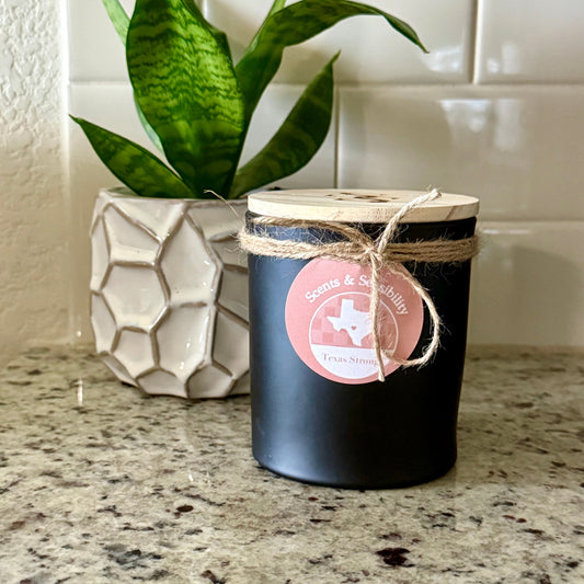 Black candle with a pink label on a marble countertop next to a potted plant.
