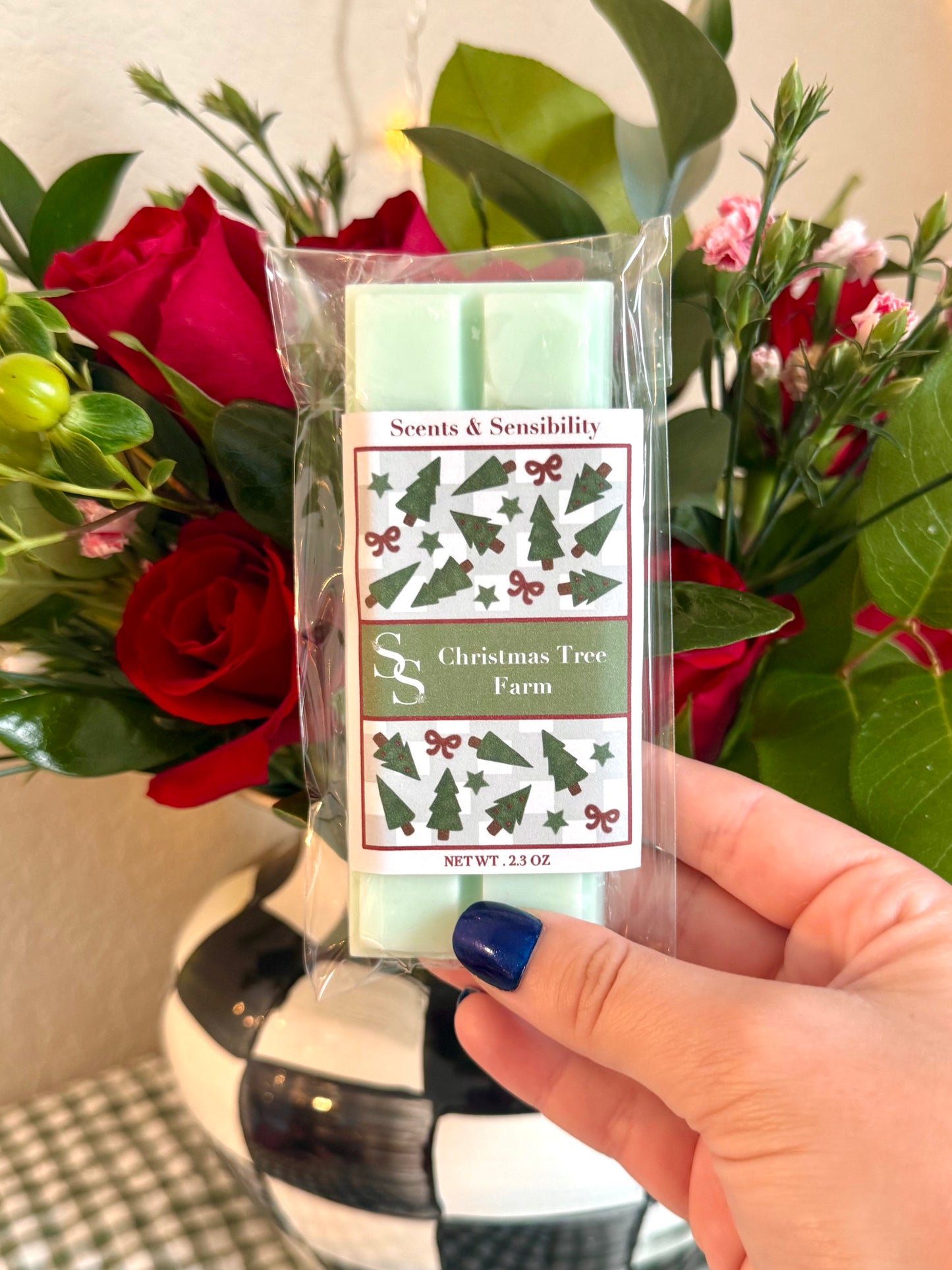 Hand holding a Scents & Sensibility candle labeled 'Christmas Tree Farm' with flowers in the background.